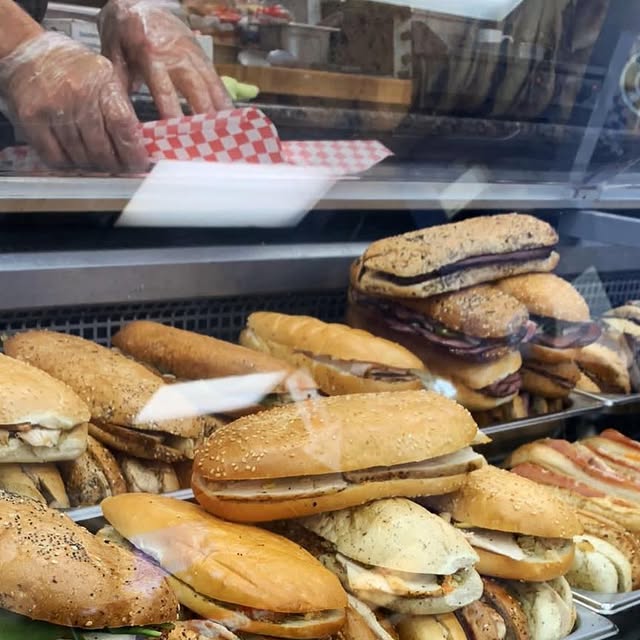 Hands preparing fresh paninis behind the glass deli case, placing sandwiches in checkered paper
